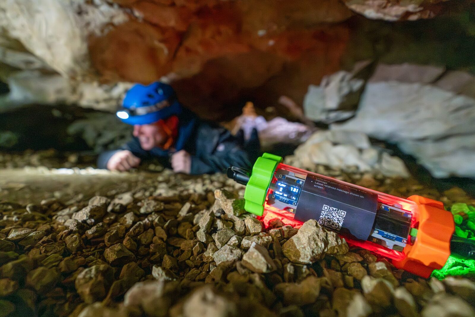 A LoCave node lying on the gravel floor of a cave, its display lit and a caver in a helmet crawling towards it in the background.